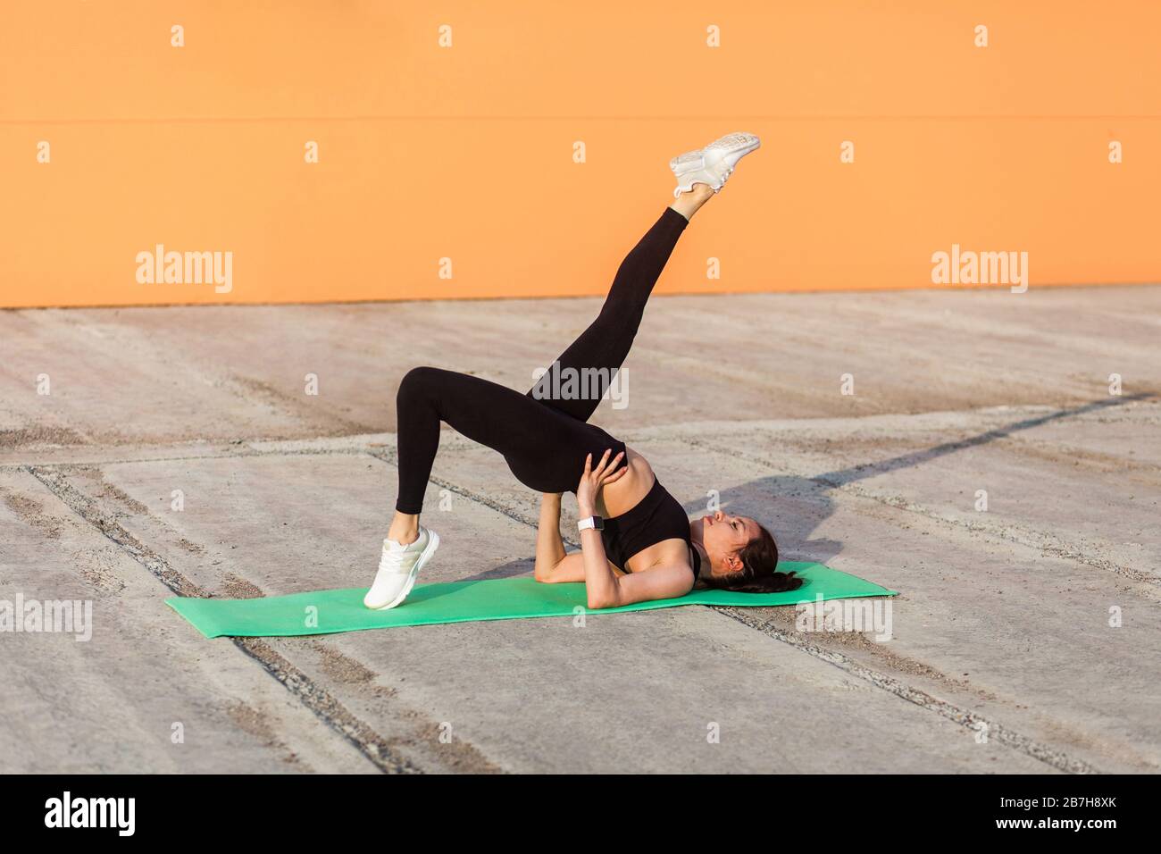 Schlankes, sportliches Mädchen in enger Sportswear, schwarzer Hose und Oberteil, praktizierendes Yoga, einbeinige Brückenpose mit Beinaufhebung, Trainingsmuskeln und Flexibilität Stockfoto