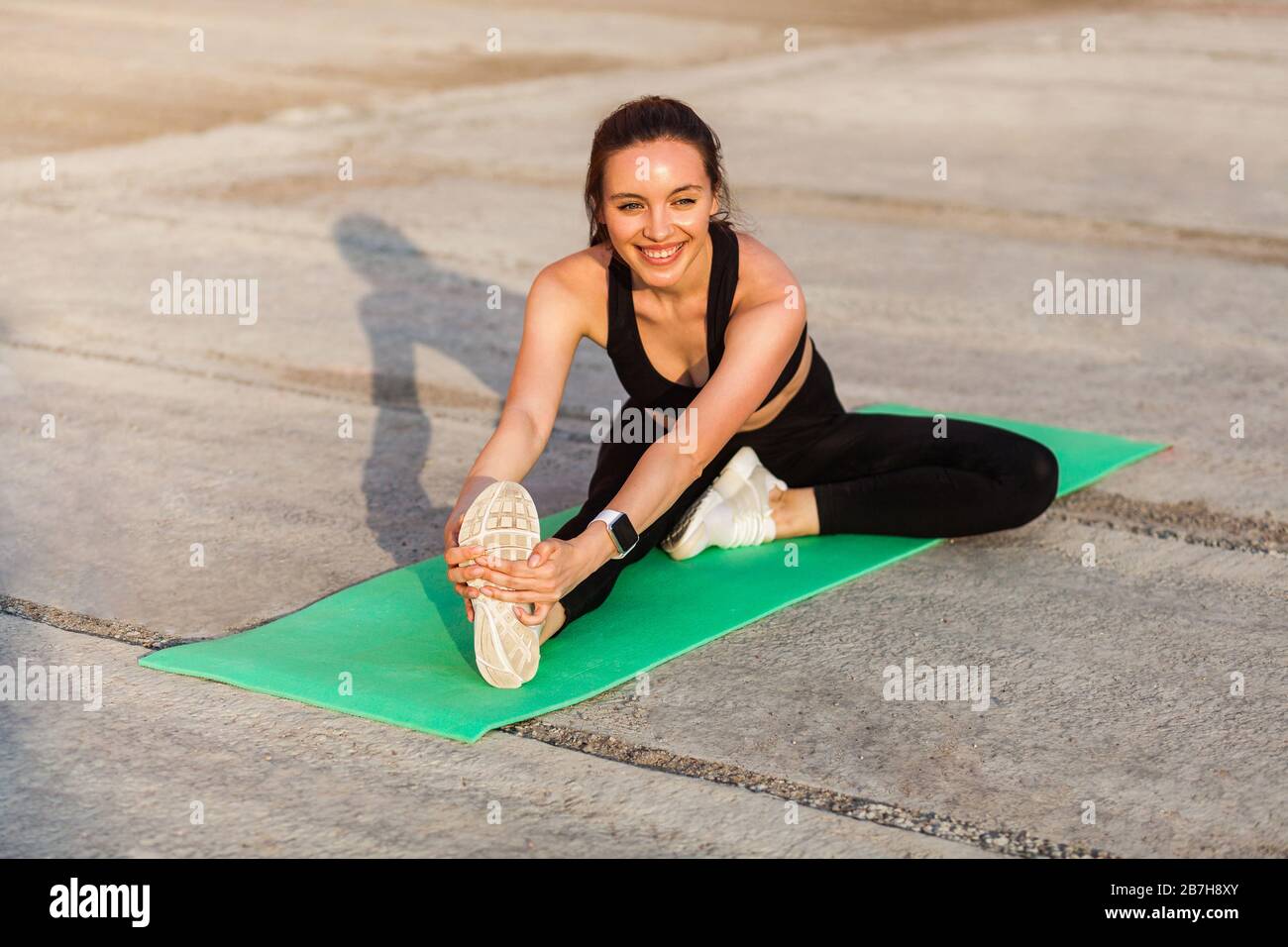 Fröhliche lächelnde Sportfrau in enger Sportswear, schwarzer Hose und Oberteil, praktizierendes Yoga, Kopf-Knie-Pose, berührende Zehen, ausdehnendes Bein und Stockfoto