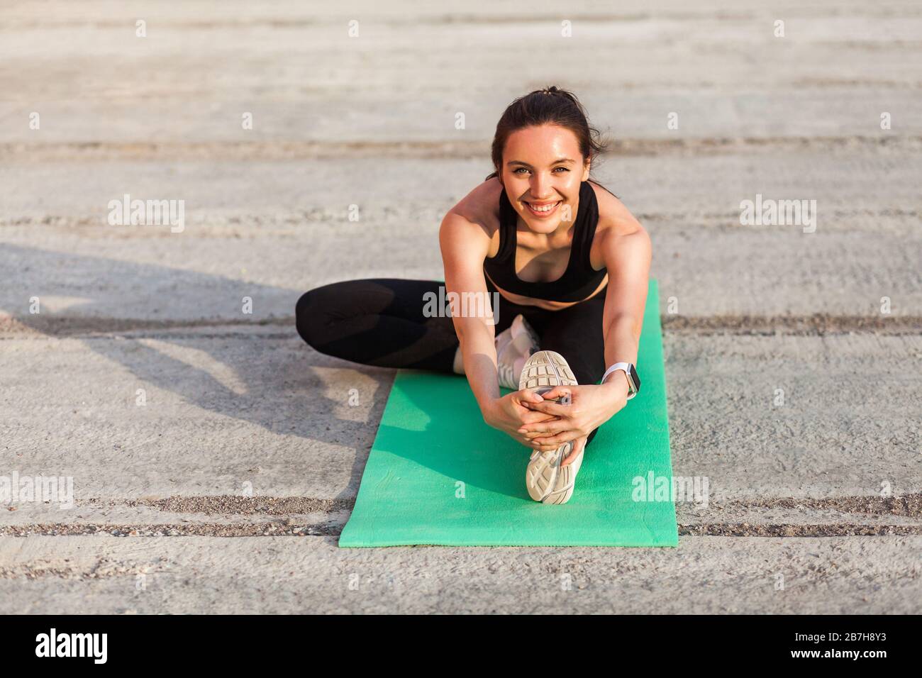 Fröhliche lächelnde Sportfrau in enger Sportswear, schwarzer Hose und Oberteil, praktizierendes Yoga, Kopf-Knie-Pose, berührende Zehen, ausdehnendes Bein und Stockfoto