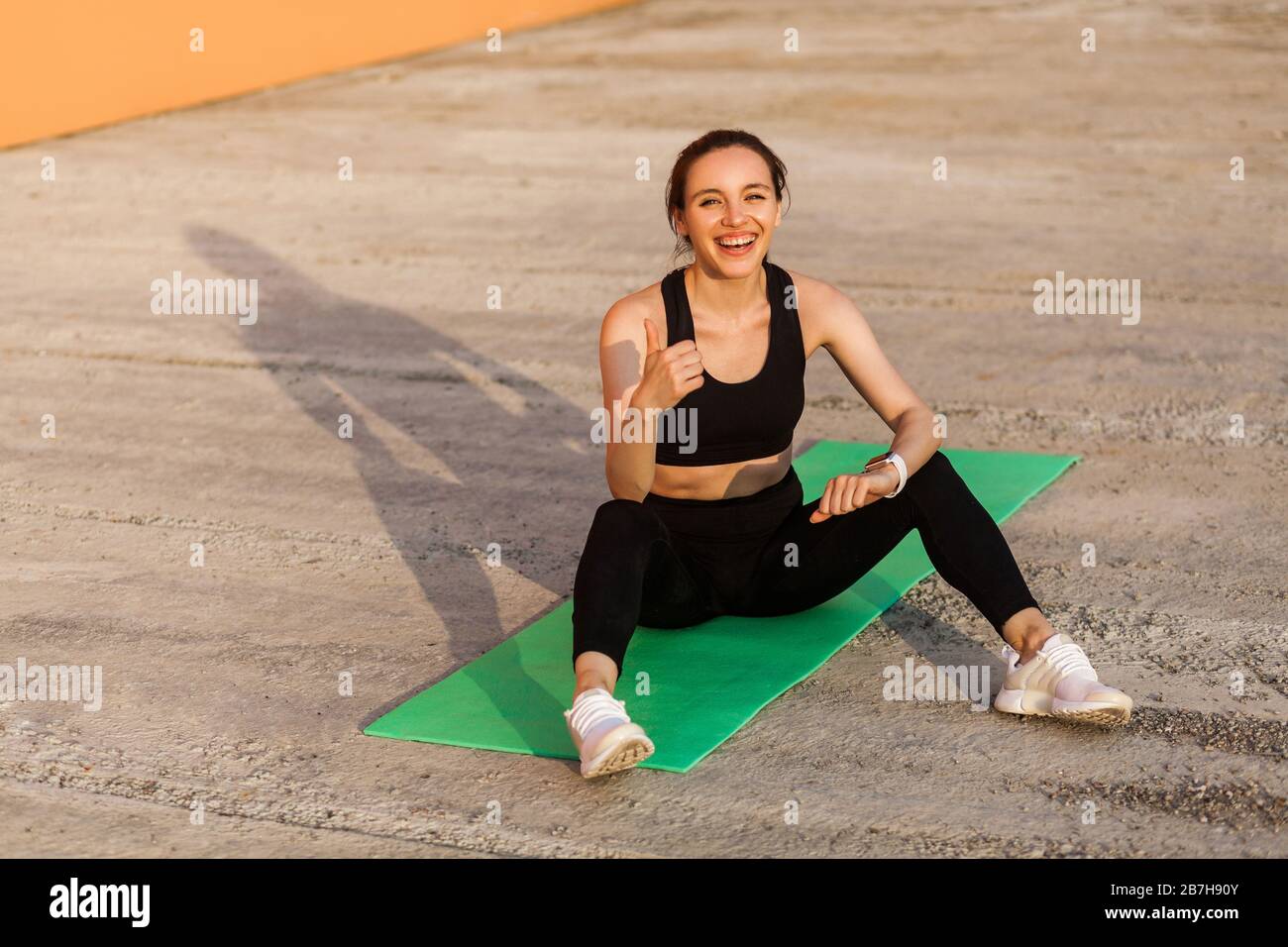 Entzücktes sportliches Mädchen in schwarzer Hose und Oberteil, lächelnd und mit Daumen nach oben, auf Yogamatte sitzend, gestikulierend wie, zufrieden mit Phys Stockfoto