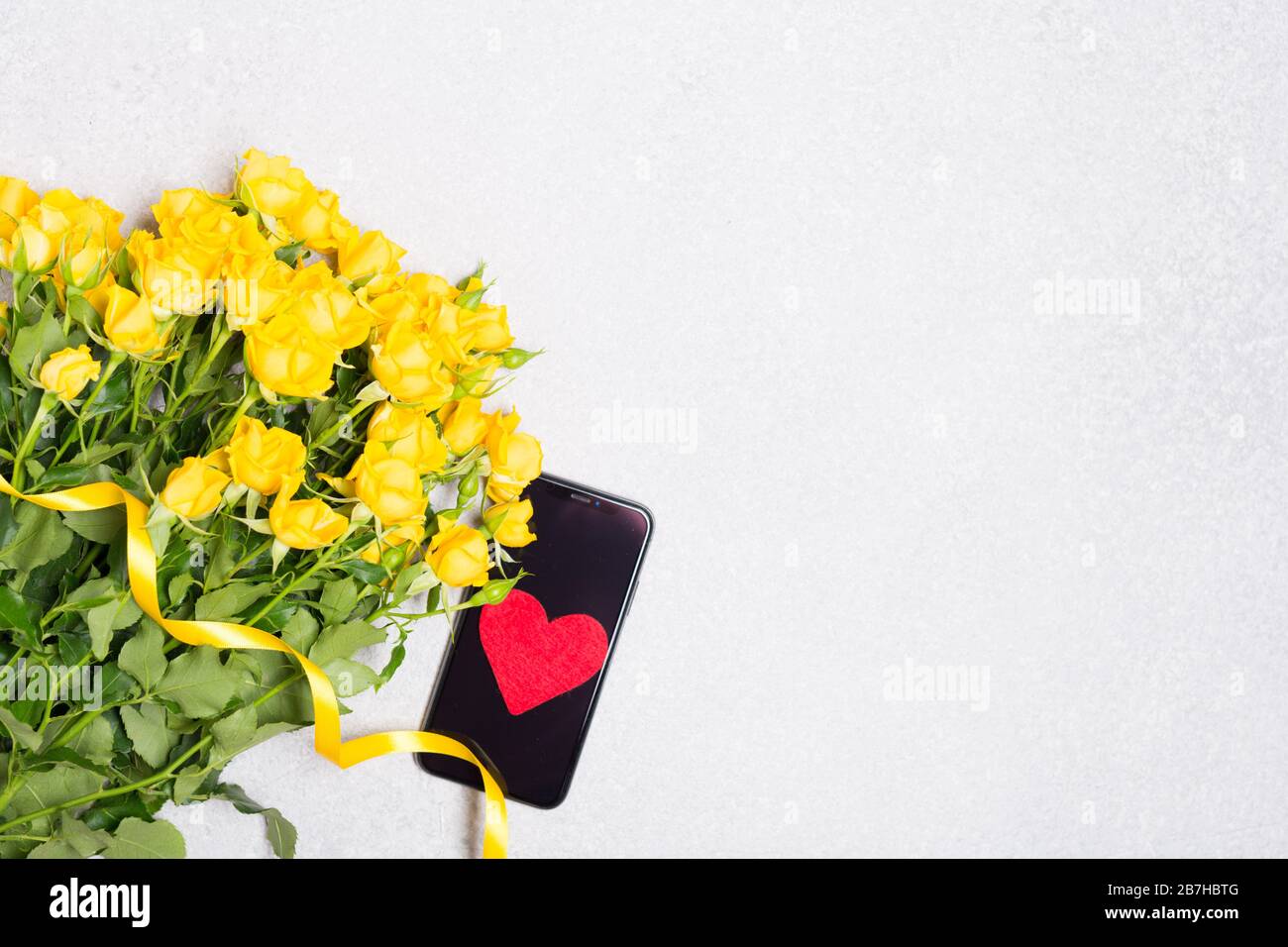 Gelbe Rosen Blumen und Telefon mit rotem Herz auf weißem Tisch Hintergrund Draufsicht verspottet freien Platz Stockfoto
