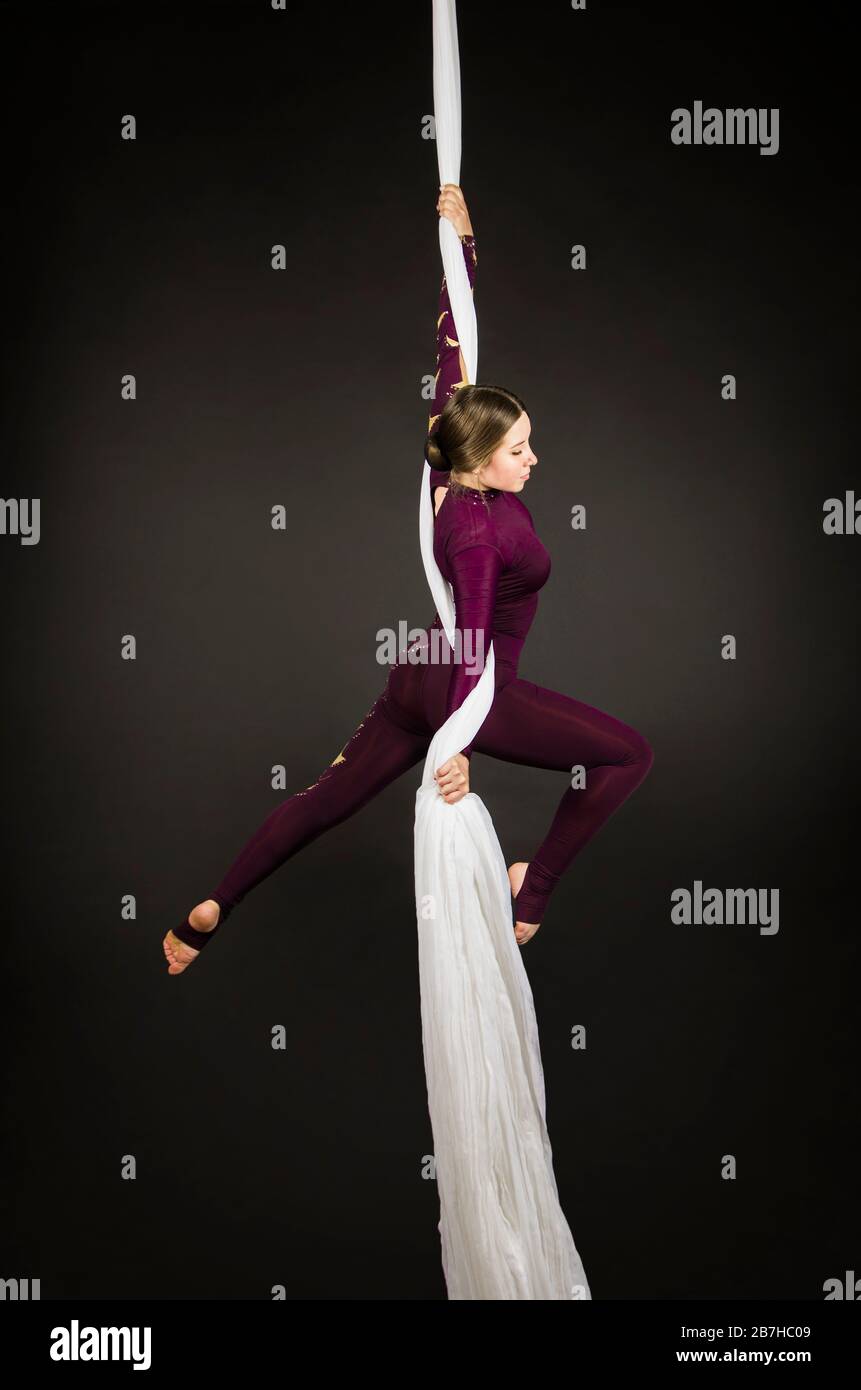 Sportliche Frau im Burgunder Anzug führt Gymnastik- und Zirkusübungen auf weißer Seide durch. Studio-Aufnahmen vor dunklem Hintergrund, isolierte Bilder. Stockfoto