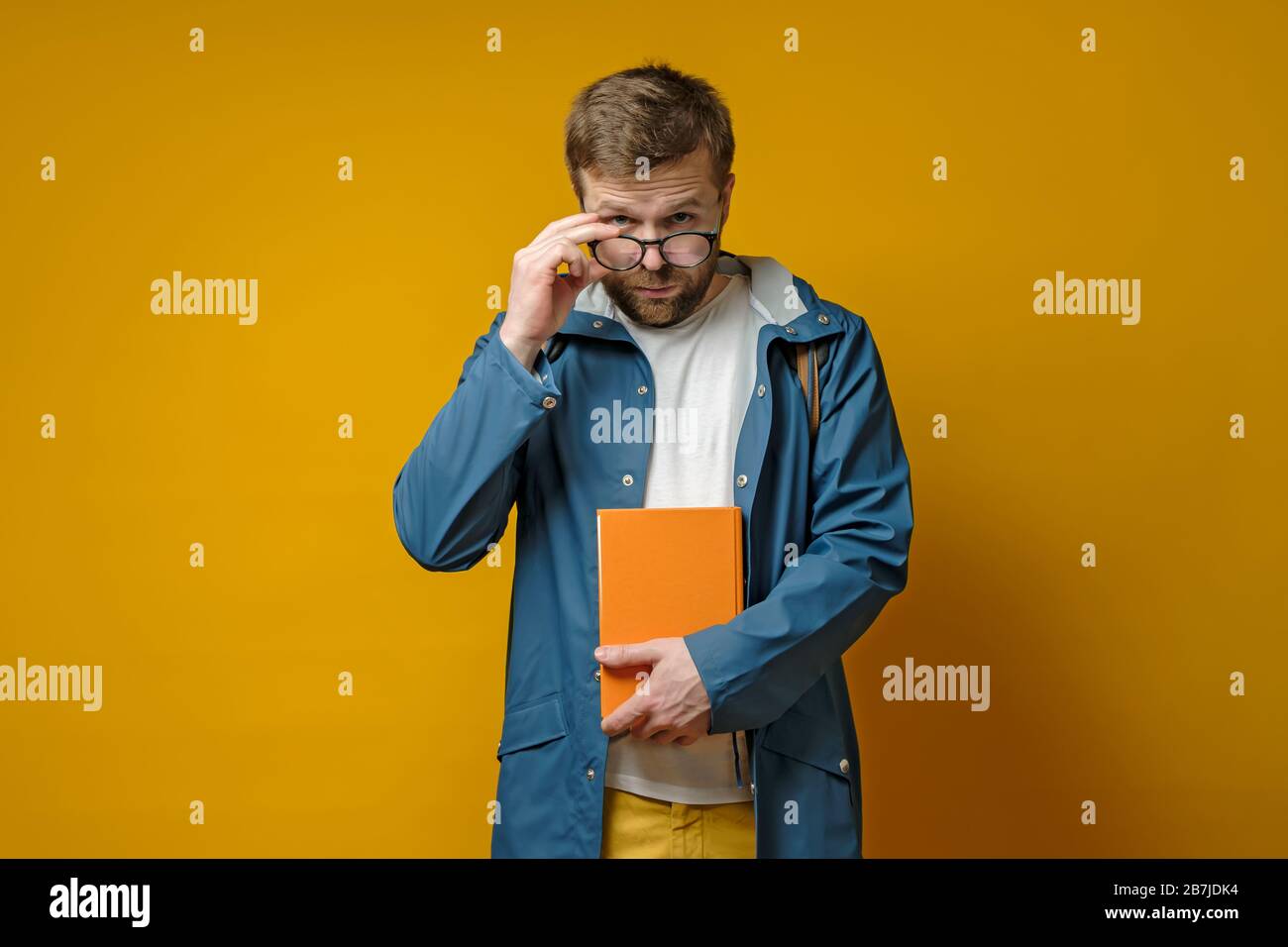 Student oder junger Wissenschaftler in einem Regenmantel und mit einem Buch, hält Brille in der Hand und sieht selbstbewusst aus, auf gelbem Hintergrund. Stockfoto
