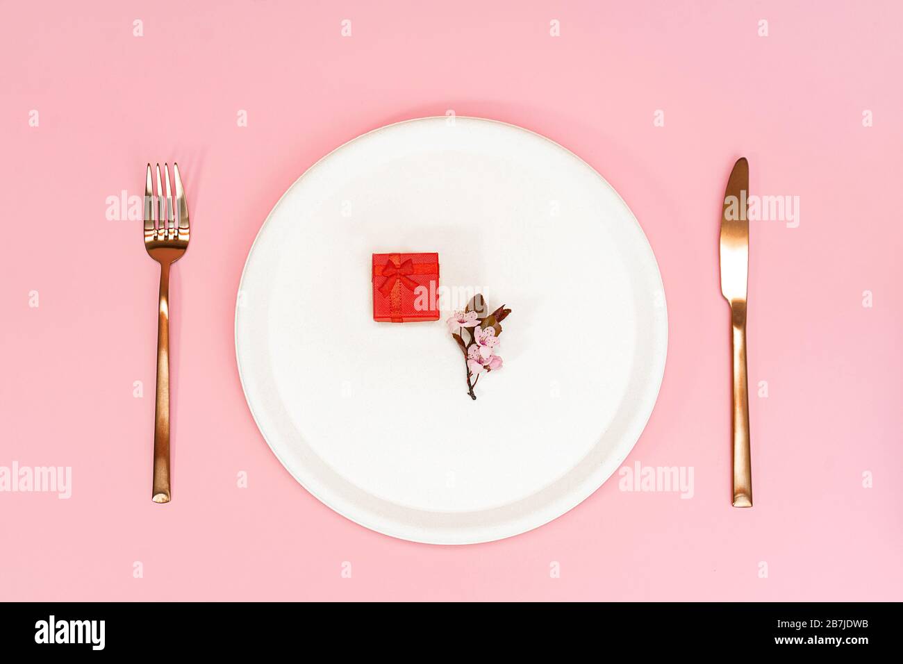 Frühlingskirschblüte und roter Geschenkkasten auf cremefarbendem Teller, Messer und Gabel. Auf pinkfarbenem Hintergrund. Flaches Layout, Kopierbereich. Kirschblüte. Stockfoto