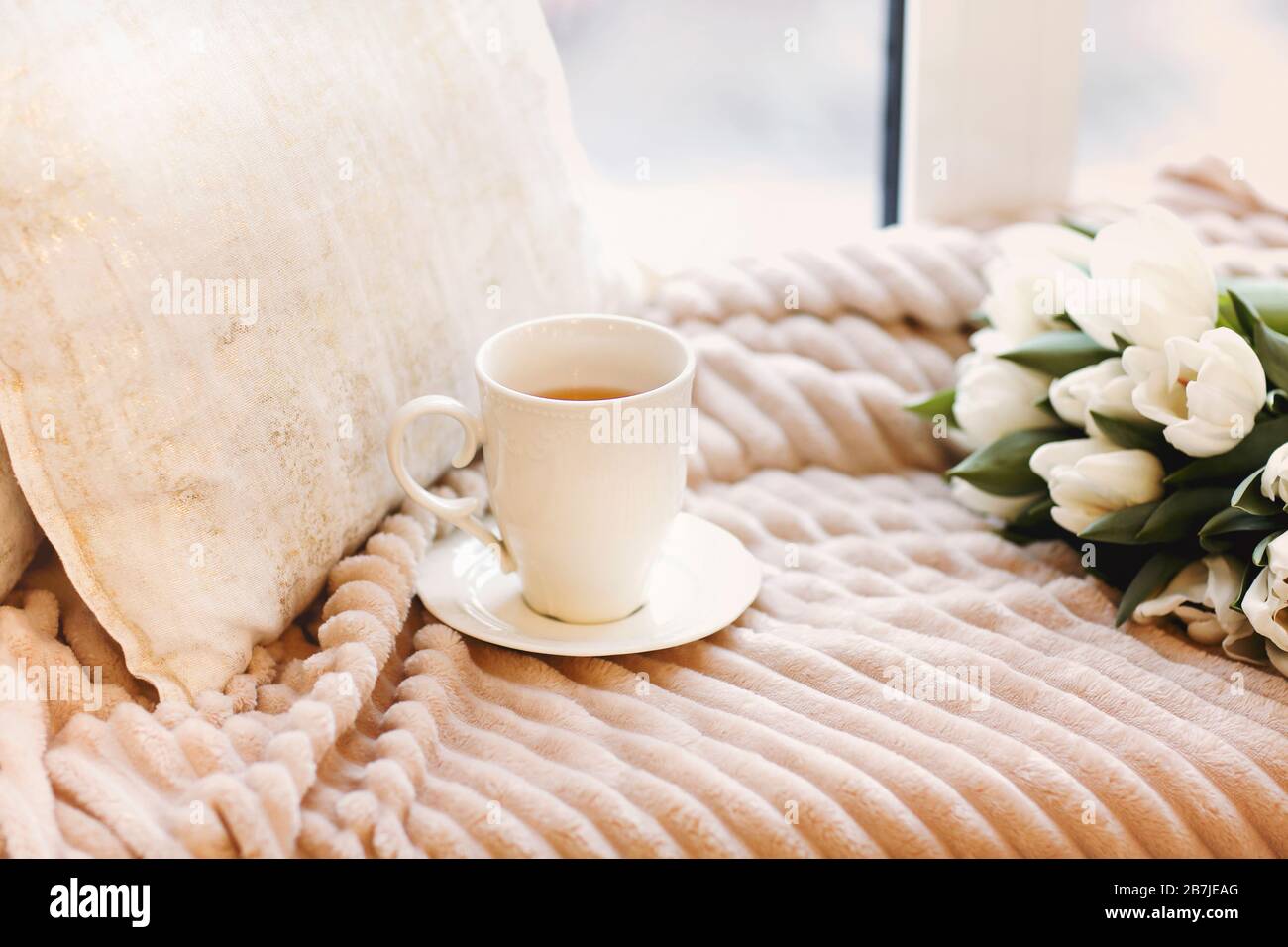 Blumenstrauß aus frischen weißen Tulpen und weißer Tasse Kaffee auf einem gemütlichen Bett mit beigefarbener Plüschdecke und Kissen im hellen Schlafzimmer Stockfoto