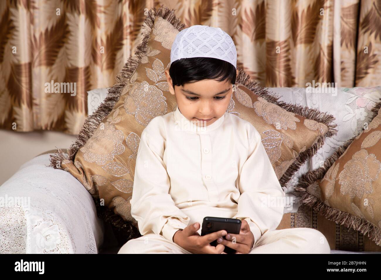 Muslimisches Kind, das zu Hause ein Mobiltelefon verwendet, während es auf dem Sofa sitzt Stockfoto