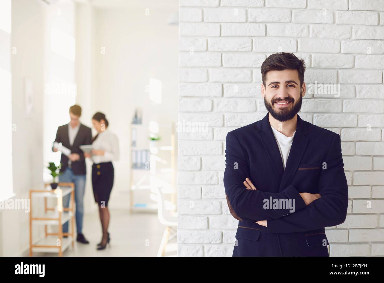 Positiver, glücklicher bärtiger Geschäftsmann in einer Jacke lächelnd, während er vor einem weißen Bürohintergrund steht. Stockfoto