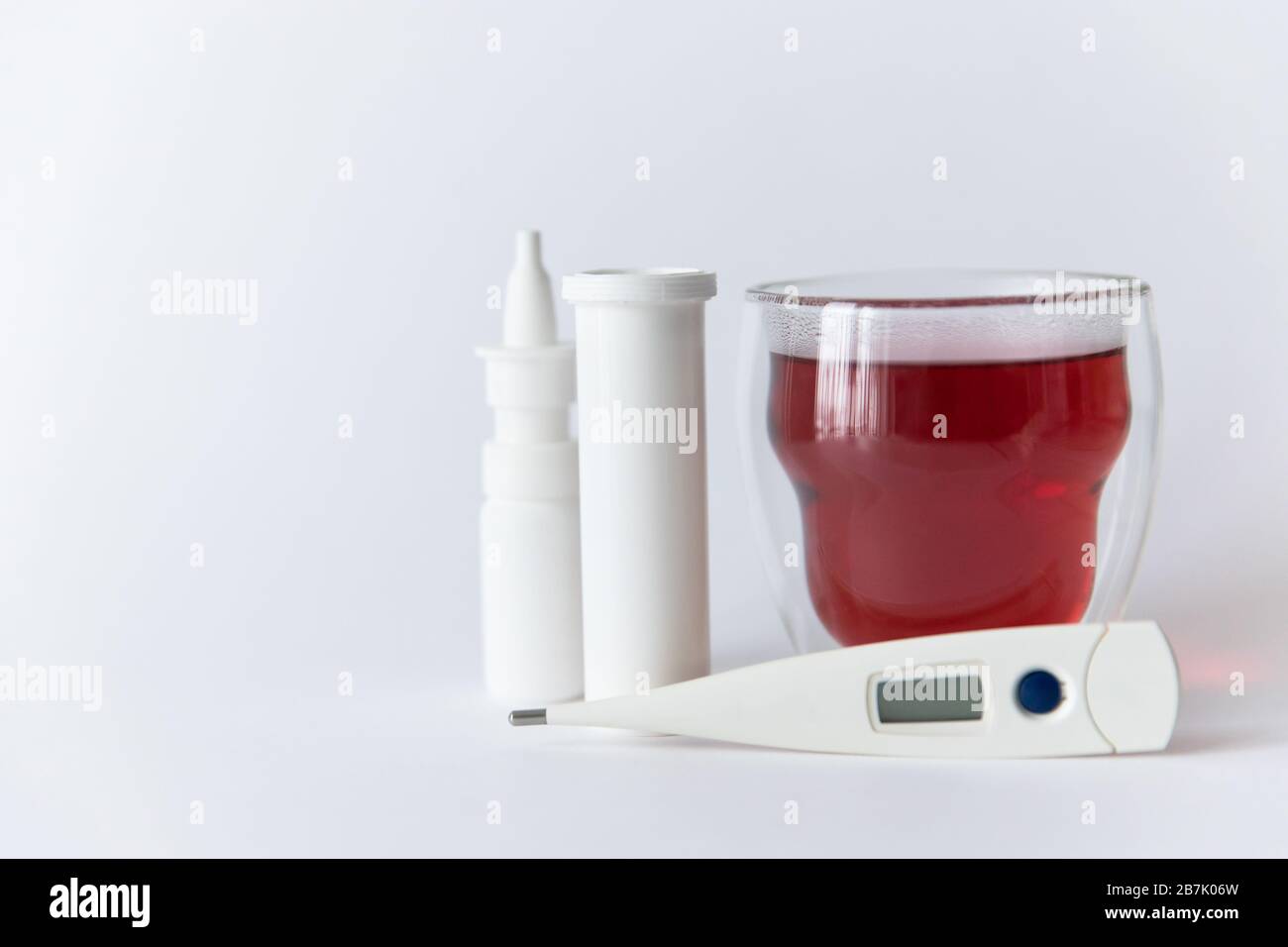 Nahaufnahme der nasalen Spritzmedizin, der weißen Pillenflasche, des medizinischen Digitalthermometers und des Glases mit rotem heißem Tee, isoliert auf weißem Hintergrund mit Kopiespac Stockfoto