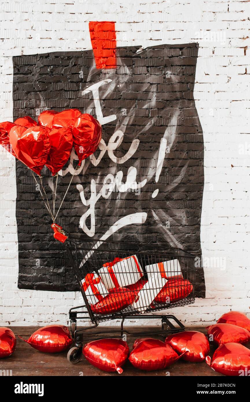 Weiße Mauer aus Backstein mit Graffiti, die ich liebe, ein schwarzer Lebensmittelkorb aus Metall mit Geschenkschachteln mit roten Schleifen und Foil Hearts Ballons überall. Stockfoto