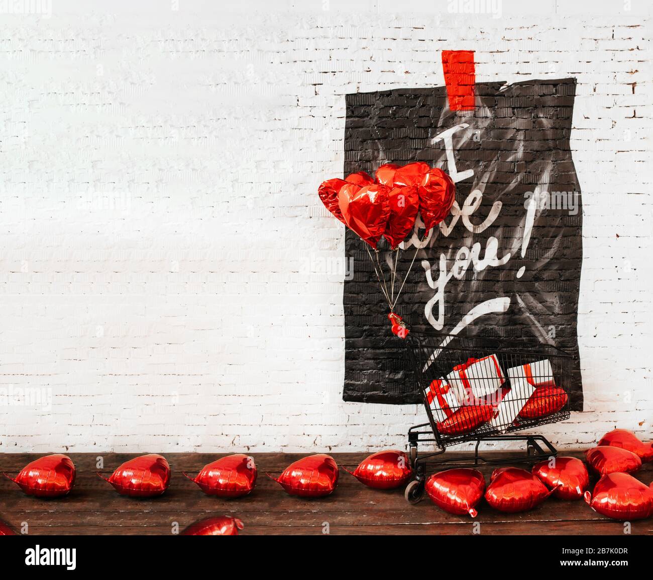 Weiße Mauer aus Backstein mit Graffiti, die ich liebe, ein schwarzer Lebensmittelkorb aus Metall mit Geschenkschachteln mit roten Schleifen und Foil Hearts Ballons überall. Layout Stockfoto