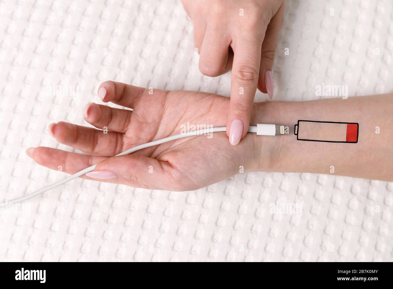 Symbol für niedrigen Batteriestand auf der menschlichen Hand. Müde Frau, die auf dem Bett liegt und versucht, das Gerät über USB aufzuladen und das Kabel mit dem Finger zu halten. Überarbeit, erschöpft, Stockfoto