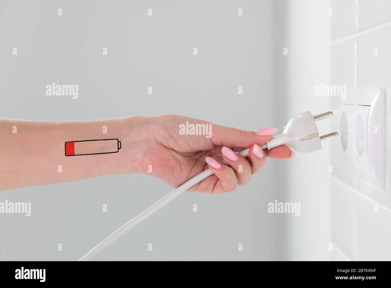 Symbol für niedrigen Batteriestand an Hand/Handgelenk des Menschen. Müde Frau hält weißen Stecker und erreicht Steckbuchse. Überarbeit, erschöpft, chronische Ermüdung Stockfoto