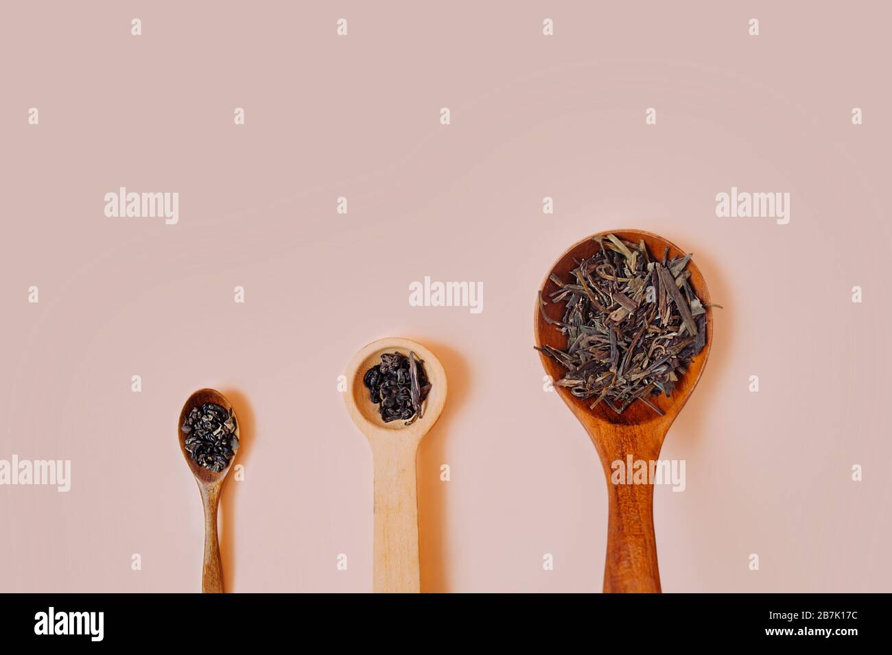 Drei Vintage-Löffel aus Holz in verschiedenen Größen mit trockenem Granulat und getrockneten Teeblättern auf einem zarten pinkfarbenen Pfirsich-Hintergrund. Stockfoto