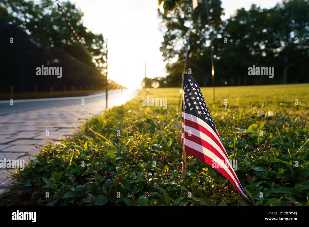 Vietnam Veterans Memorial US Flag Washington DC // WASHINGTON DC — Eine kleine US-Flagge wird im Gras vor dem Vietnam Veterans Memorial in Washington DC gepflanzt und fängt die Morgensonne ein. Das Vietnam Veterans Memorial, entworfen von Maya Lin, beleuchtet durch das sanfte Licht des Sonnenaufgangs. Die Wände aus schwarzem Granit mit den Namen gefallener Soldaten spiegeln den frühen Morgenhimmel wider und schaffen eine feierliche und reflektierende Atmosphäre in der National Mall. Stockfoto