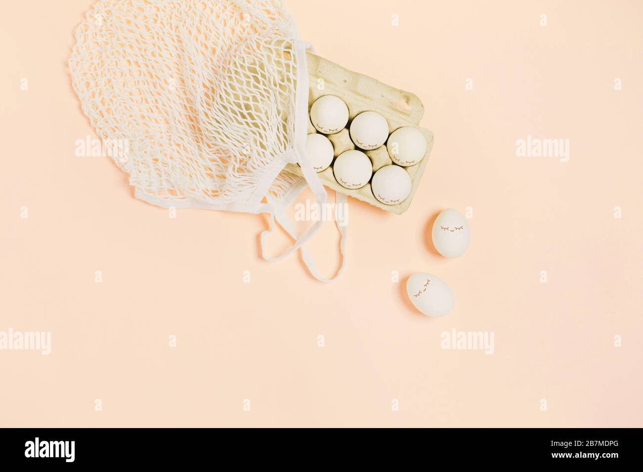 Frohe Ostern. Weiße Eier mit bemalten Augen in Papierverpackungen und weißer Netztasche auf Pfirsichfarbenhintergrund. Flaches Layout, Draufsicht, Kopierbereich. Stockfoto