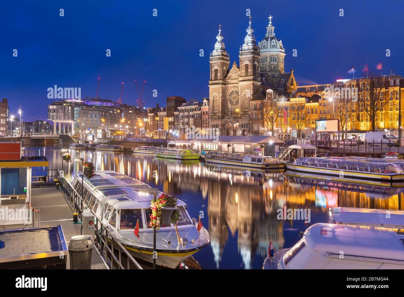 Amsterdam, Niederlande, Kanalszene in der Nacht mit der Basilika Sankt Nikolaus und Flussbooten. Stockfoto