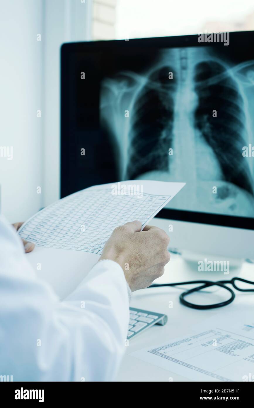 Nahaufnahme eines jungen Arztes, der einen weißen Mantel trägt, an seinem Schreibtisch sitzt und die Ergebnisse eines Tests und eines Röntgenstrahlers in der Brust in seinem Computer überprüft Stockfoto