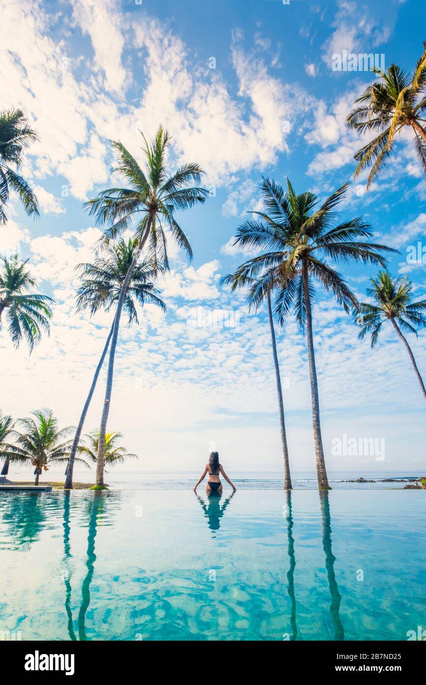 Eine junge Frau, die unter Palmen in einem Infinity-Pool steht Stockfoto