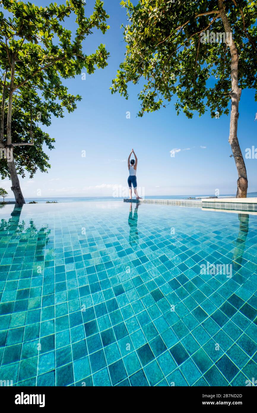 Eine junge Frau, die Yoga unter Palmen neben einem Infinity-Pool praktiziert Stockfoto