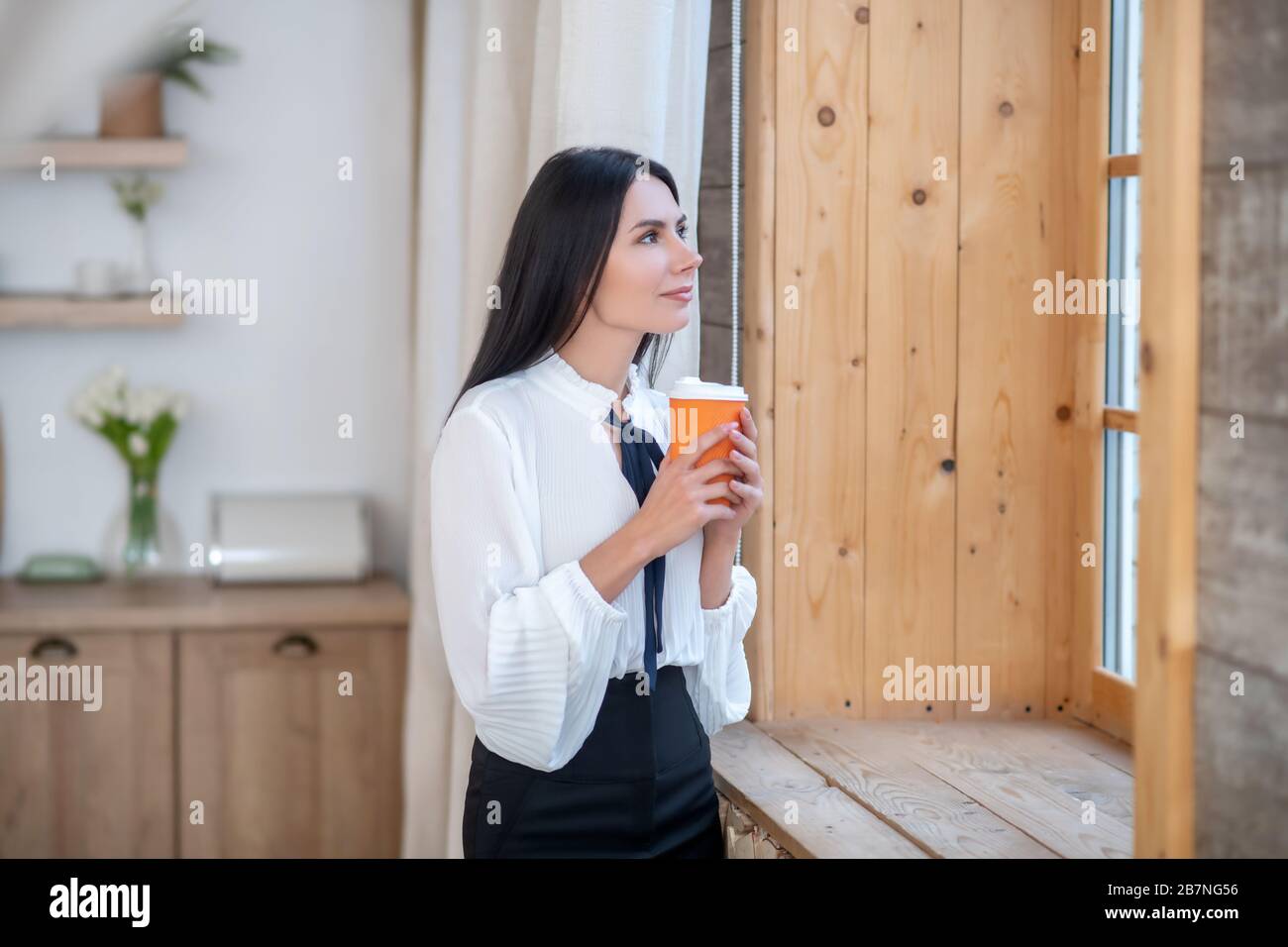 Junge Frau, die am Fenster steht und die Kaffeetasse hält Stockfoto