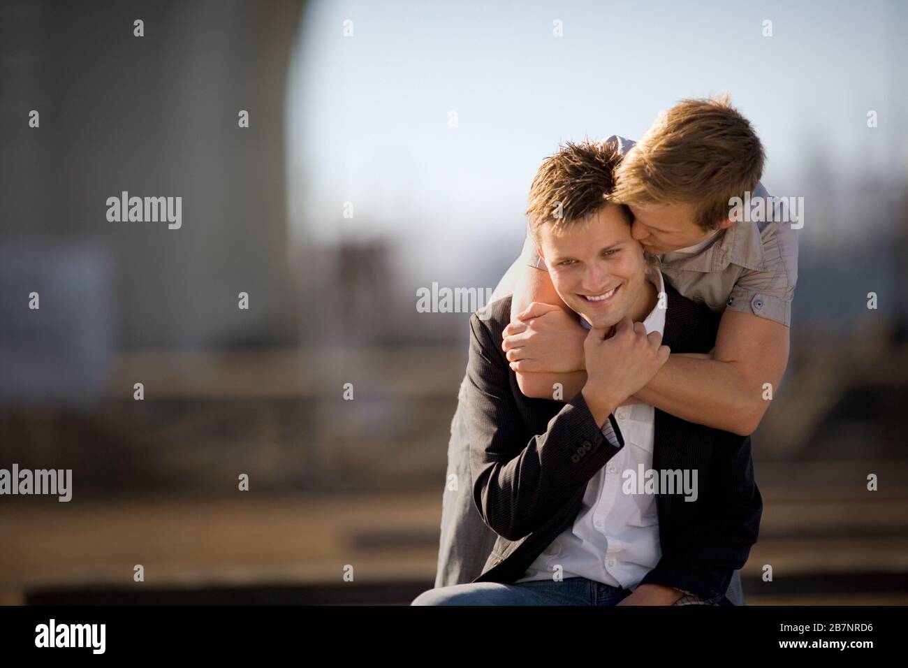 Der junge Mann, der seinem Freund einen Kuss auf das Ohr gibt, während er mit seinen Armen um ihn herum steht. Stockfoto