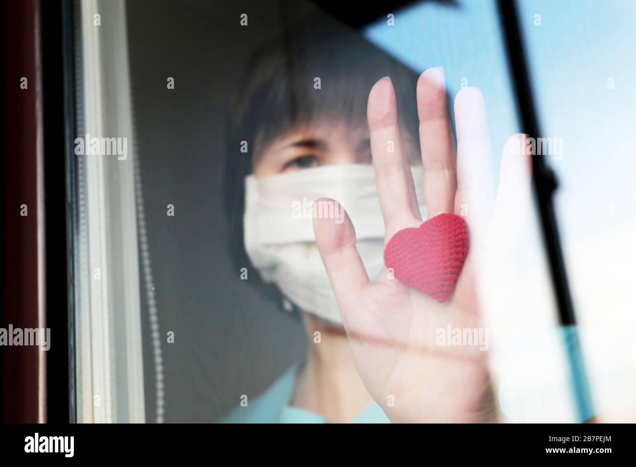 Quarantäne während der COVID-19-Coronavirus-Epidemie, infizierter Patient. Kranke Frau in einer medizinischen Gesichtsmaske blickt mit rotem Herz durch das Fensterglas Stockfoto