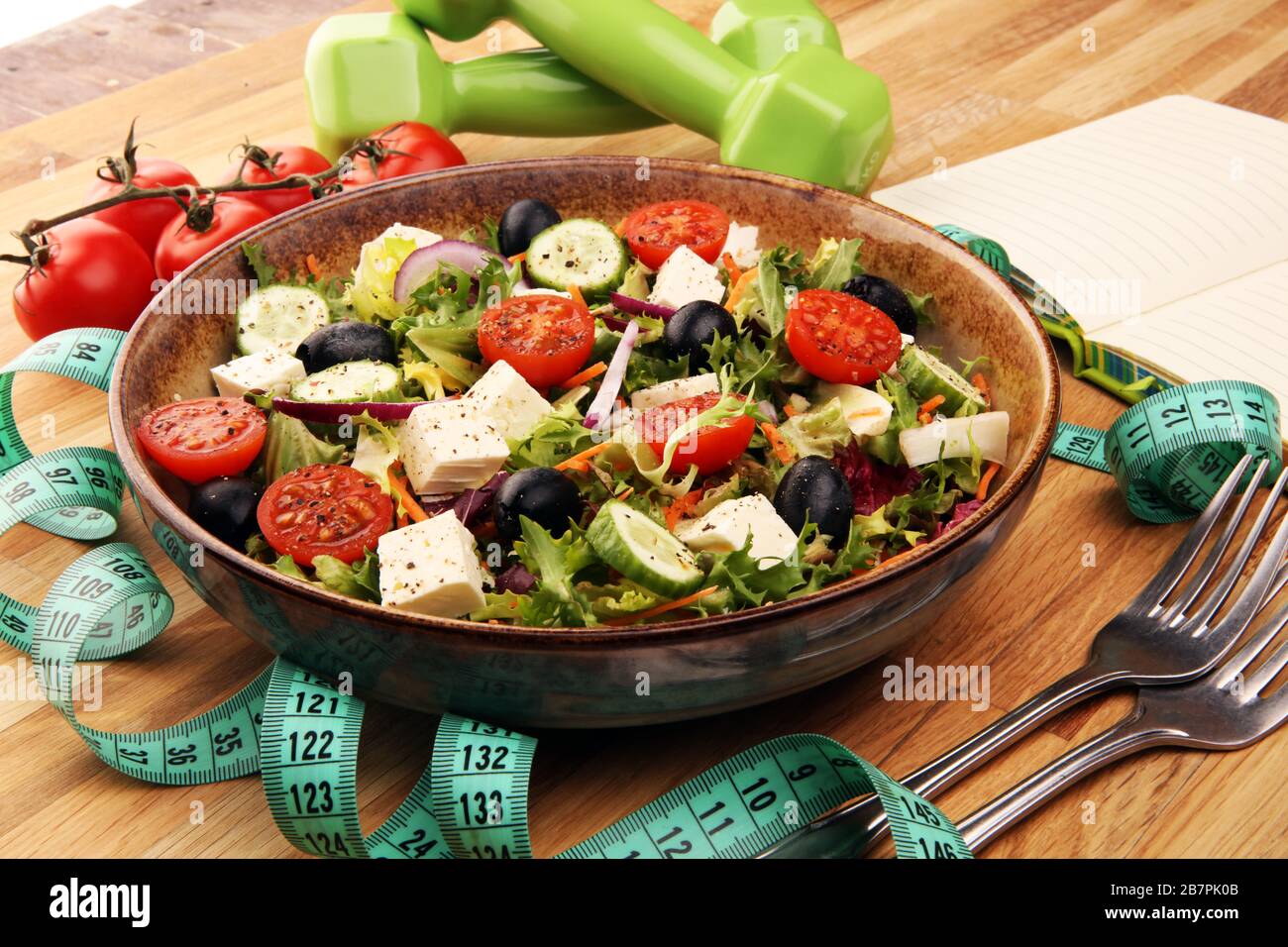 Authentischer frischer Salat in einem Steinbecher mit Hanteln Übungsausrüstung, Messband und Gabel auf dem Tisch. Gesunder Lebensstil, gutes Gesundheitskonzept. Stockfoto