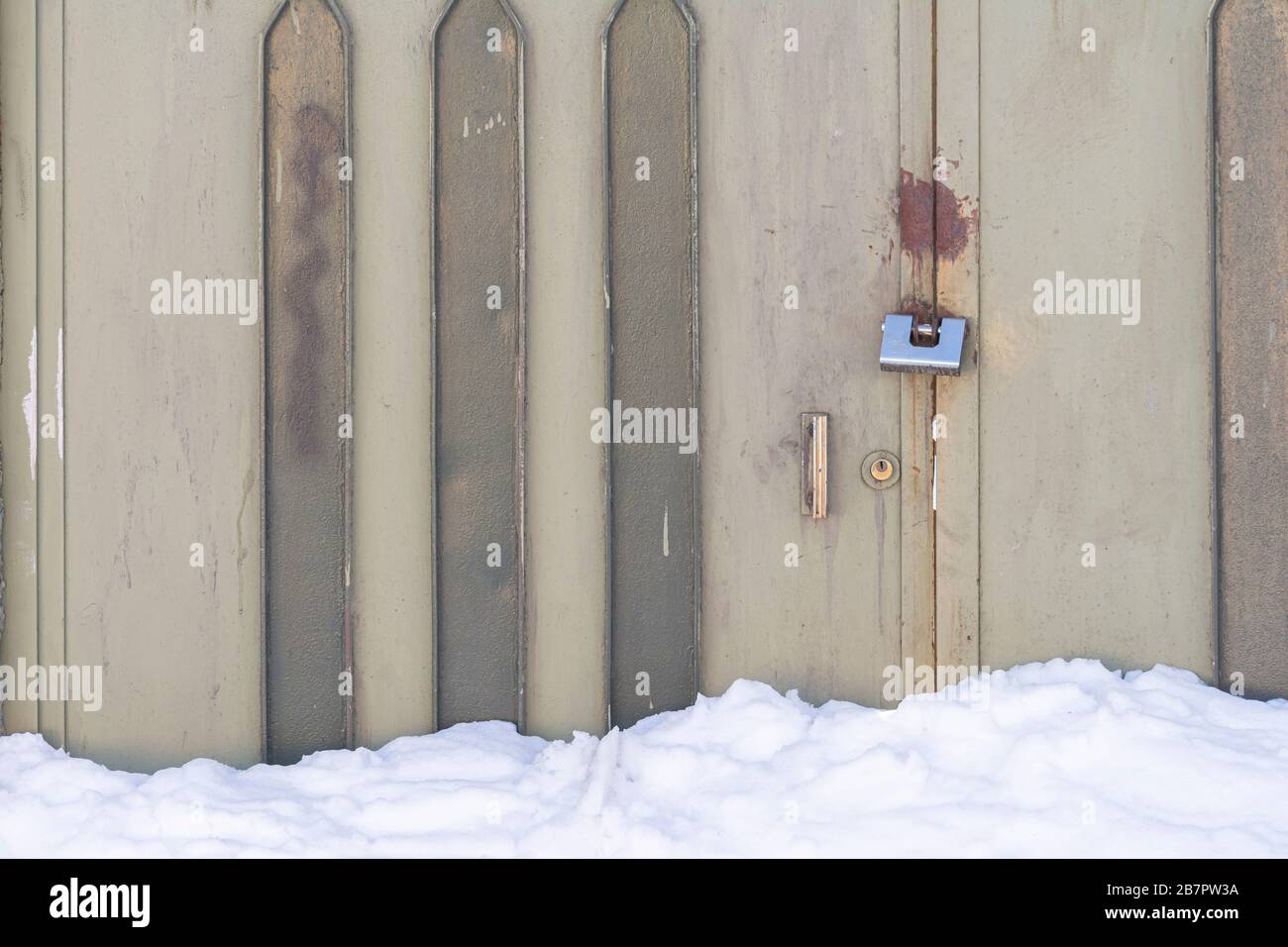 Die alte rostige Tür ist mit einem Stahl-Vorhängeschloss befestigt, vor der Tür ist Winterschnee gestapelt Stockfoto