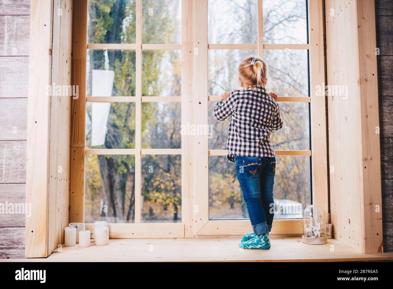 Weihnachtskonzept. Ein Kind mit blonden Haaren in warmen Socken, Jeans und Hemden steht mit dem Rücken auf dem Fensterstock und legt ihre Hände auf den Stockfoto