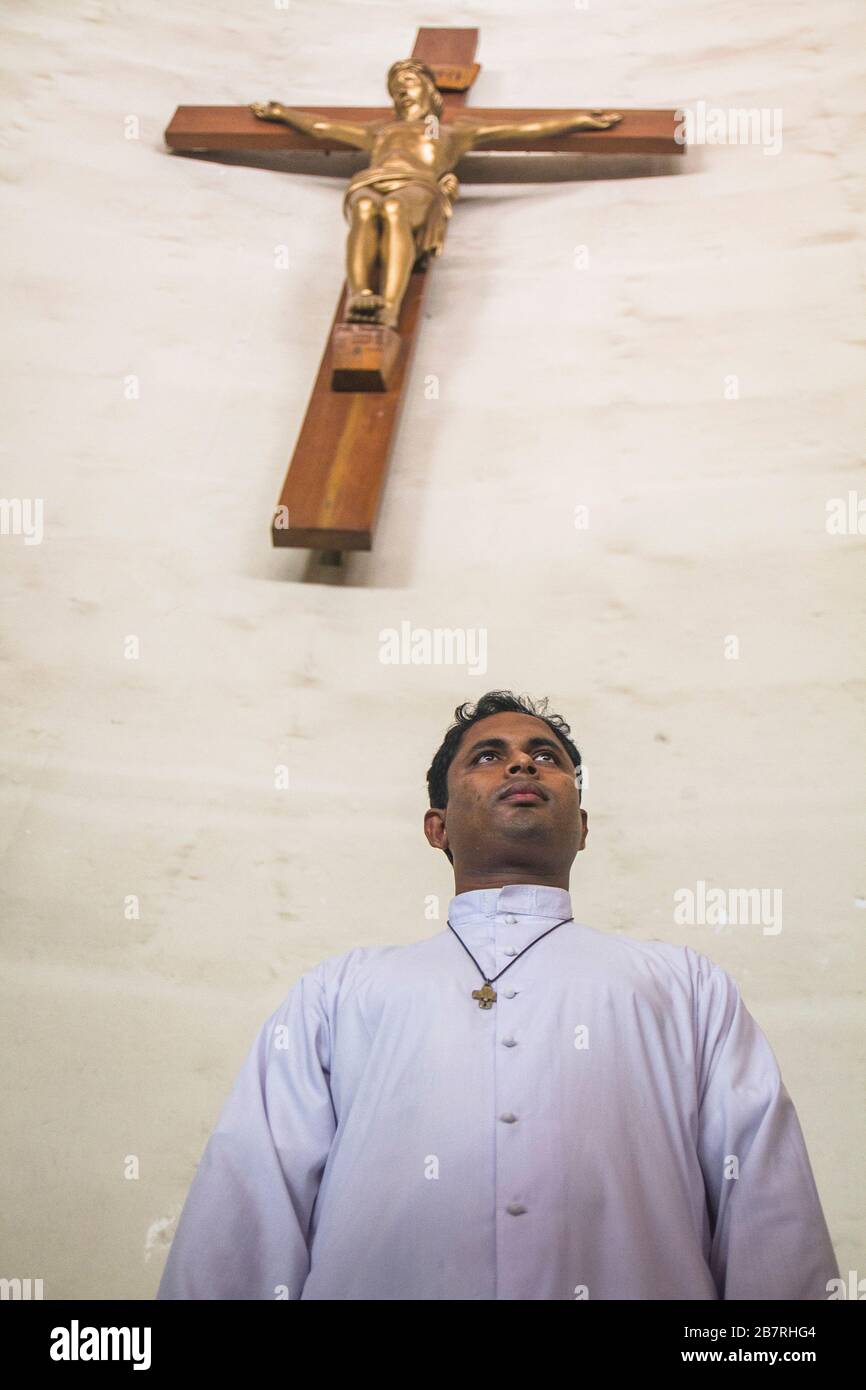 Ein Priester der Tejgaon-Kirche mit weißem Kleid, das unter einem Kreuz steht und einem Geistlichen einen Blick nach vorne gibt. Er hat gerade nur wenige Kinder getauft. Stockfoto