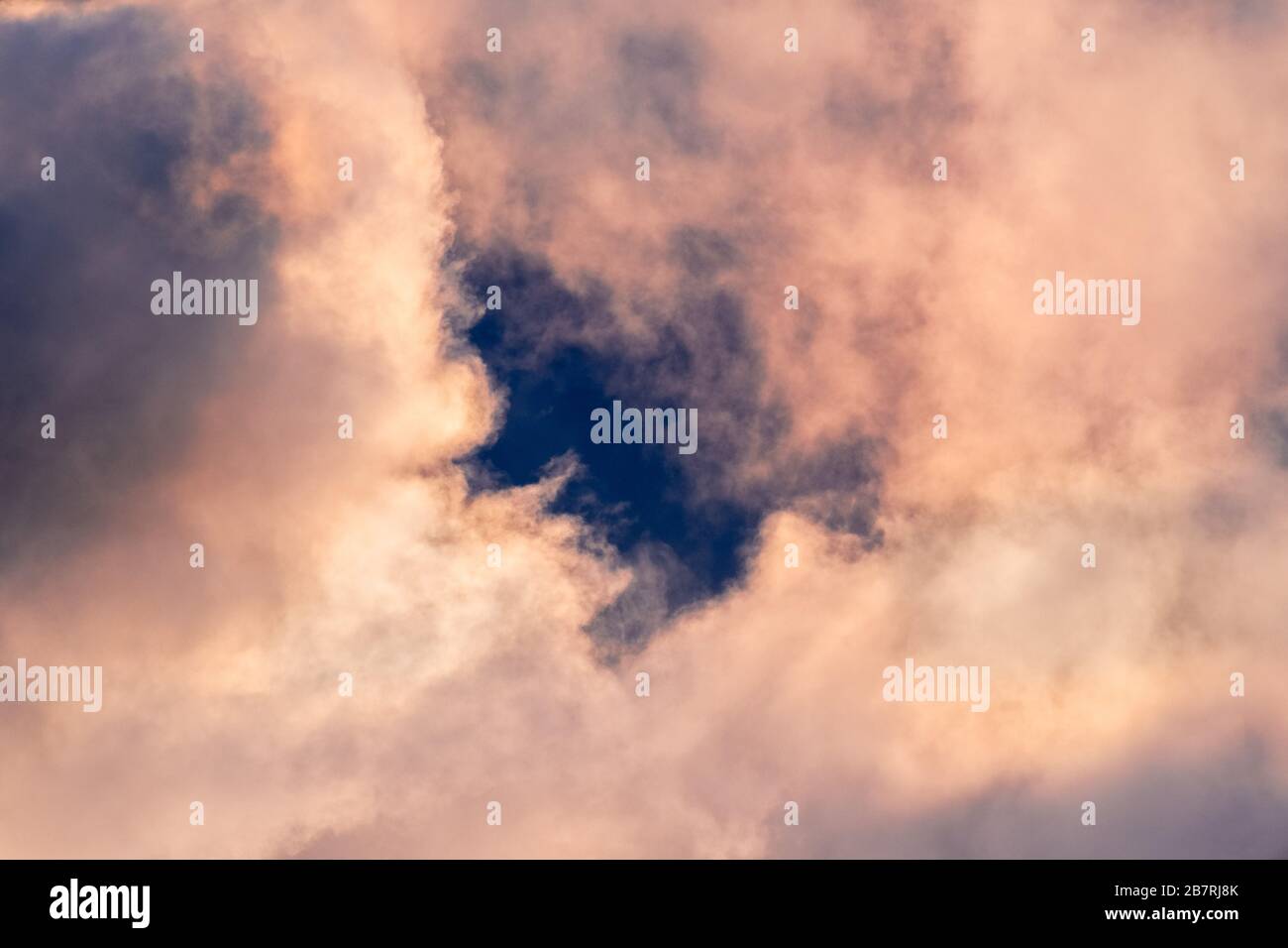 Nahaufnahme von Sturmwolken, die in Farben bei Sonnenuntergang eingefärbt sind; beim Einbrechen der Wolken wird dunkelblauer Himmel sichtbar Stockfoto