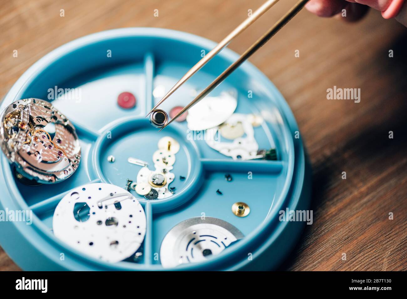 Beschnittener Blick auf den Uhrmacher, der das Uhrenteil mit Teilen in der Werkzeugablage auf dem Tisch in der Pinzette hält Stockfoto