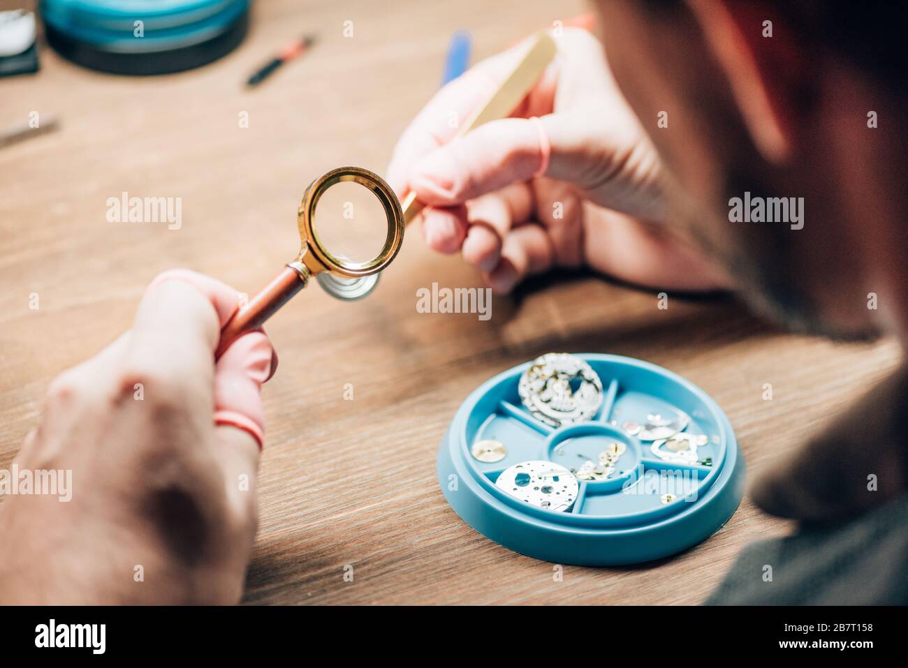 Selektive Fokussierung des Uhrmachers mit Lupe durch Werkzeugablage mit Uhrteilen auf Tisch Stockfoto