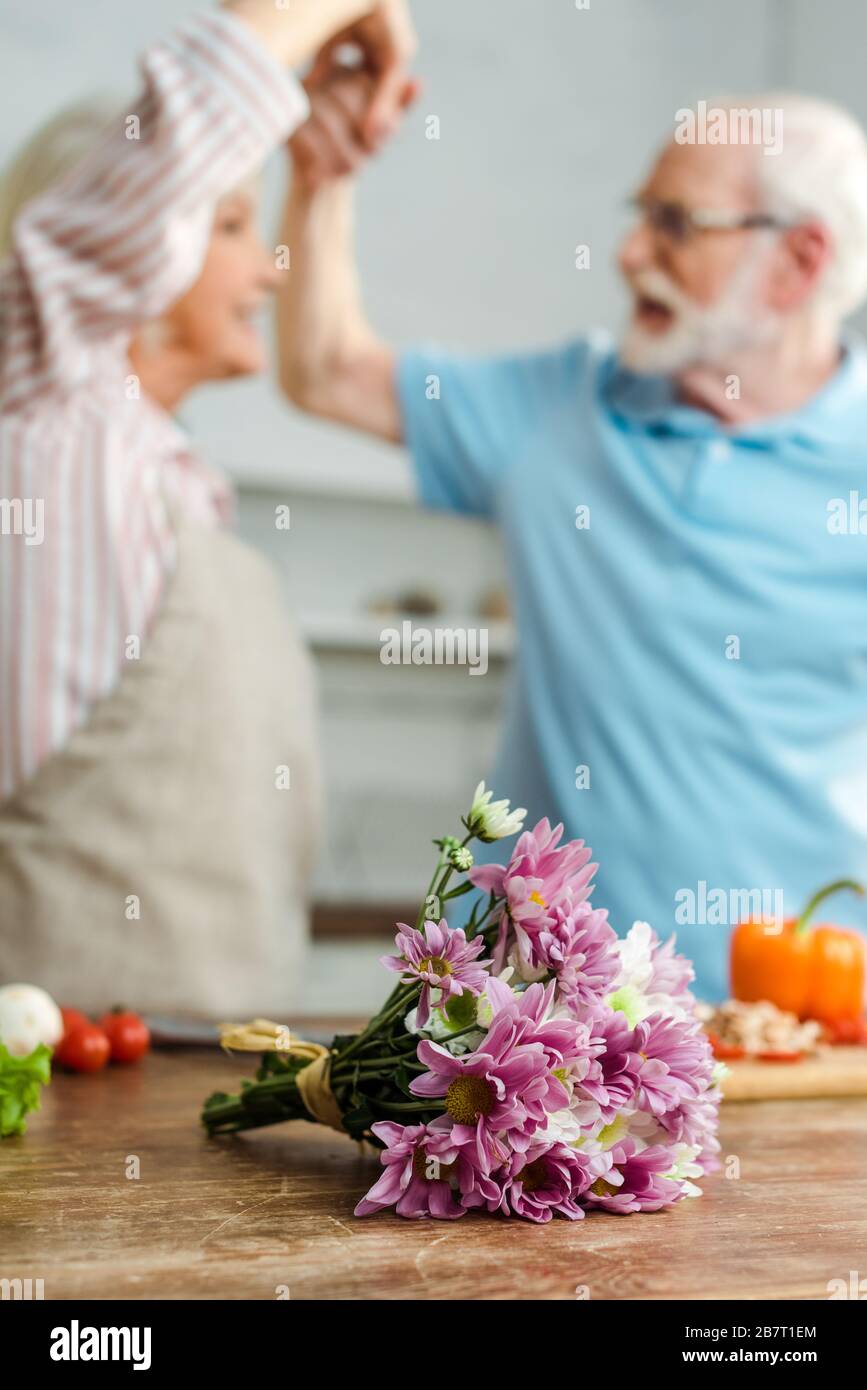 Selektiver Schwerpunkt von Blumenstrauß und Gemüse auf Küchentisch und älteres Paar, das im Hintergrund tanzt Stockfoto