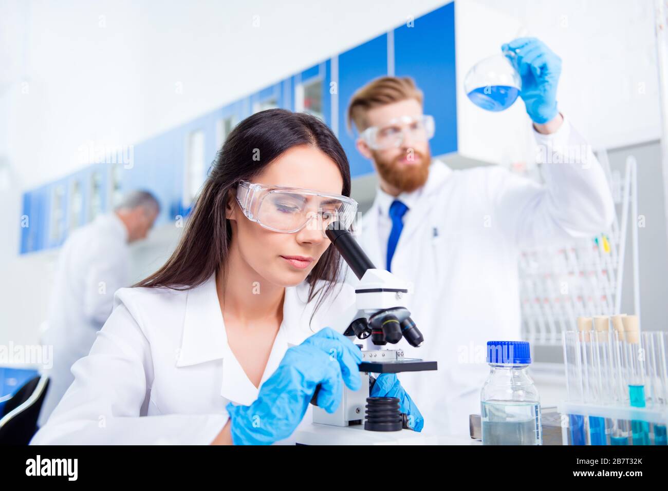 Teamarbeit. Junge Laborangestellte in Sicherheitsbrillen analysieren die Probe im Mikroskop. Sie befindet sich in einem Labcoat, mitten im Experiment Stockfoto