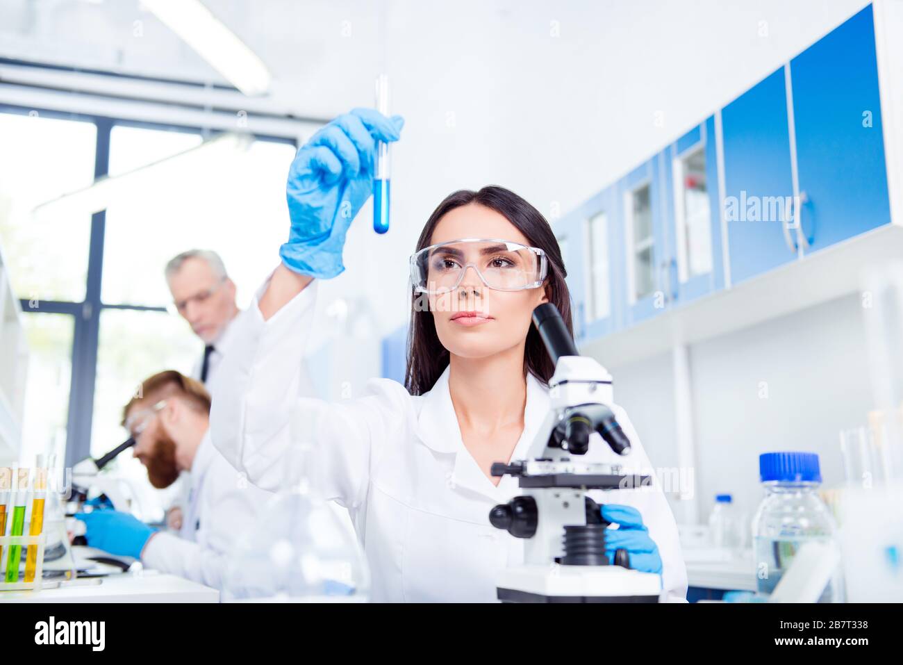 Junge brünette niedliche Laborangestellte in Sicherheitsbrillen analysieren die Probe im Laborschlauch. Sie befindet sich in einem Labcoat, mitten in der Arbeit mit dem Experiment Stockfoto