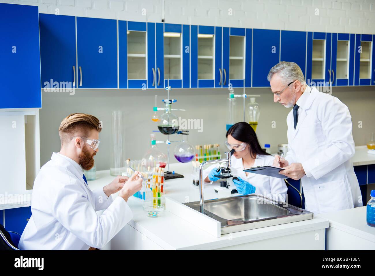 Teamwork-Konzept. Professor überprüft und steuert die Gruppenarbeit zweier Laborinterns. Alle drei konzentrieren sich auf das Experimentieren und tragen von Labcoats Stockfoto