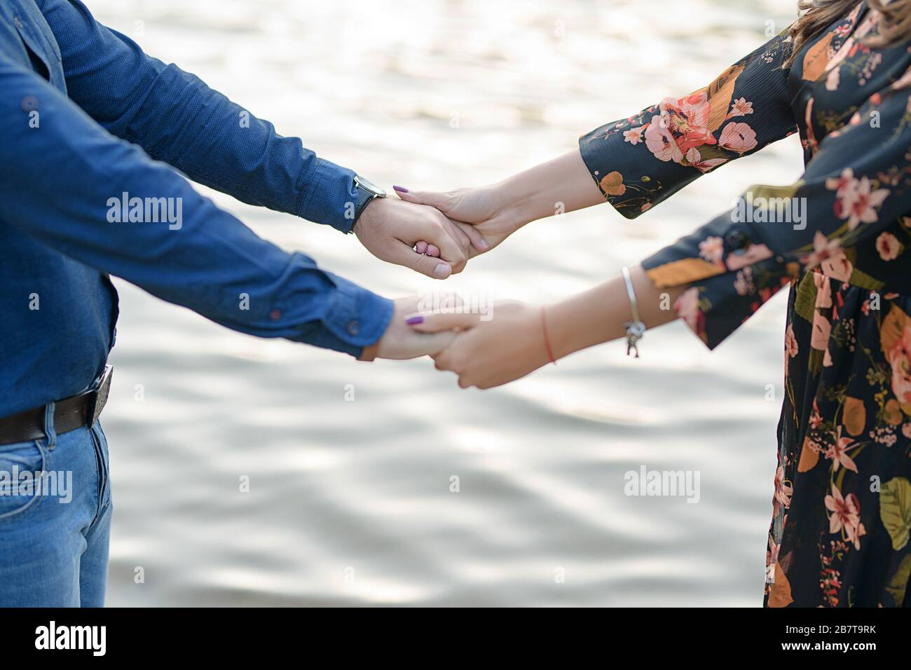 Heterosexuell kaukasisches Paar, das spielerisch die Hände vor einem unscharfen, unscharfen Wasserhintergrund hält, einem See oder einem Meer, Konzept für ein verliebtes Paar Stockfoto