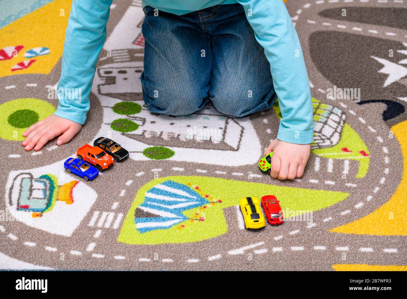 Fünf Jahre alter Junge, der auf einer Spielmatte mit Straßen Spielzeugautos spielt und auslegt. Der Junge ist in blaue Jeans gekleidet und hält einen Krankenwagen. Stockfoto