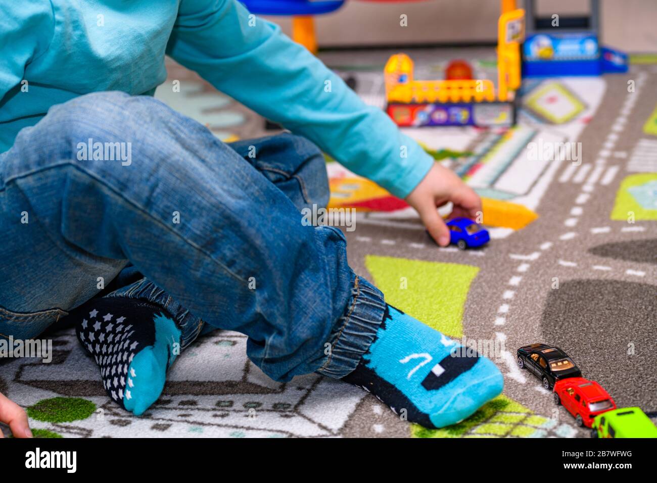 Fünf Jahre alter Junge, der auf einer Spielmatte mit Straßen Spielzeugautos spielt und auslegt. Der Junge ist mit einer blauen Jeans und einem türkisfarbenen Hemd bekleidet. Stockfoto