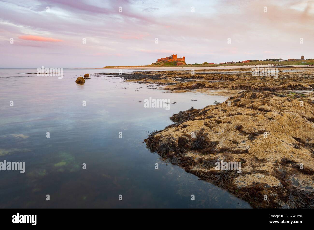 Landschaft bei Dämmerung mit Sonnenuntergang Farbe auf Himmel, Meer und die felsige Küste von Bamburgh in Northumberland mit Bamburgh Castle an der Küste in der Ferne. Stockfoto