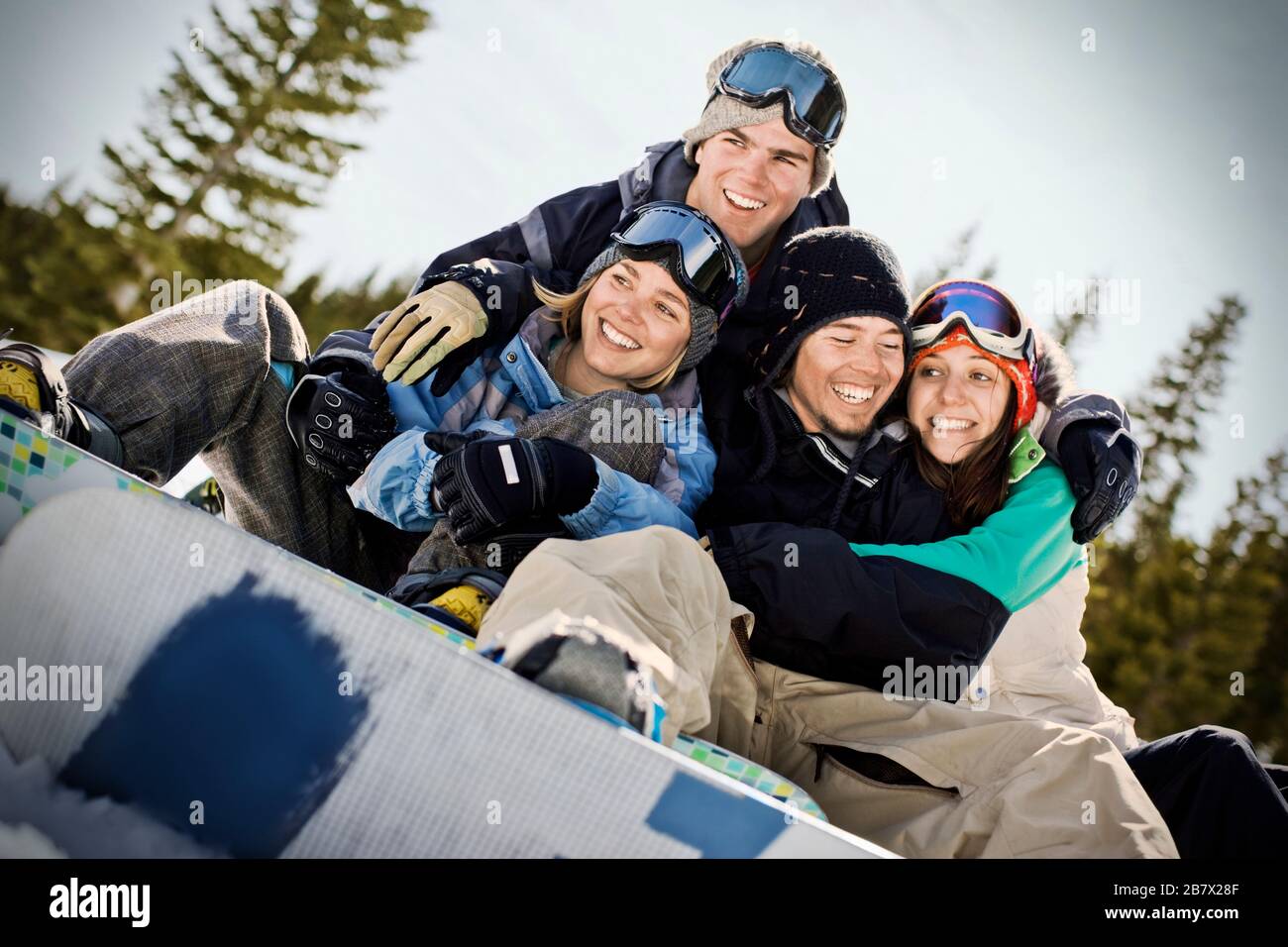 Zwei lächelnde junge Paare tragen Schneeausrüstung und sitzen mit Armen um sich herum im Freien. Stockfoto