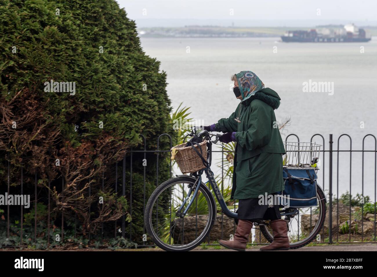Ältere Frauen tragen eine Gesichtsmaske, die ein Fahrrad entlang der Küste entlang der Steilküste über der Themse treibt, wobei ein Frachtschiff vor der Küste unterwegs ist Stockfoto