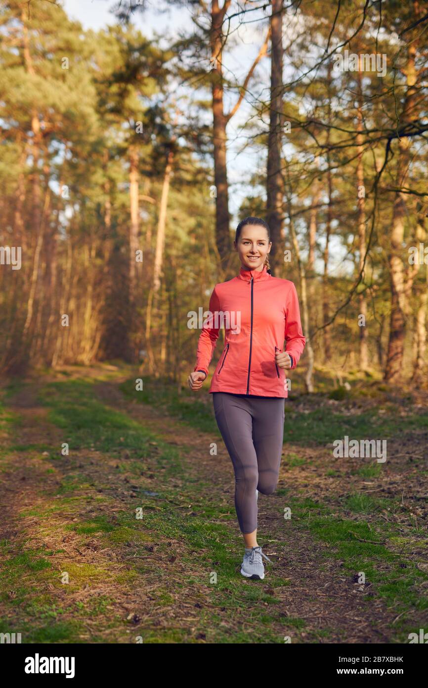 Gesunde Passform attraktive junge Frau Jogging im Wald entlang einer Waldbahn, die sich der Kamera mit einem fröhlichen Lächeln in einem aktiven Lebensstil nähert, Gesundheit a Stockfoto