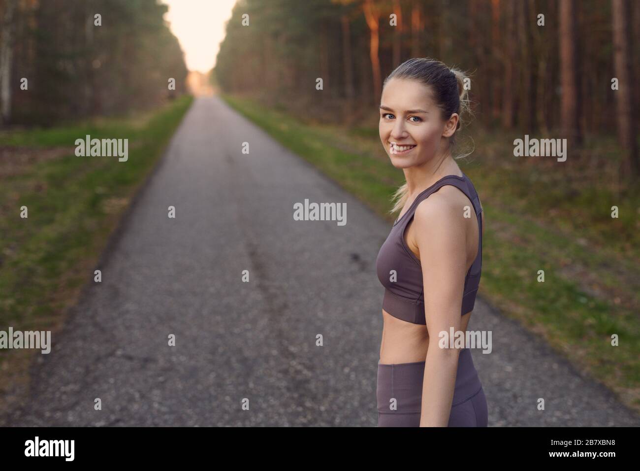 Die junge, athletische Frau passt beim Sonnenaufgang auf einer gerade sich zurückziehenden Straße durch Waldbäume und leuchtet am Ende der Sonne und lächelt Stockfoto