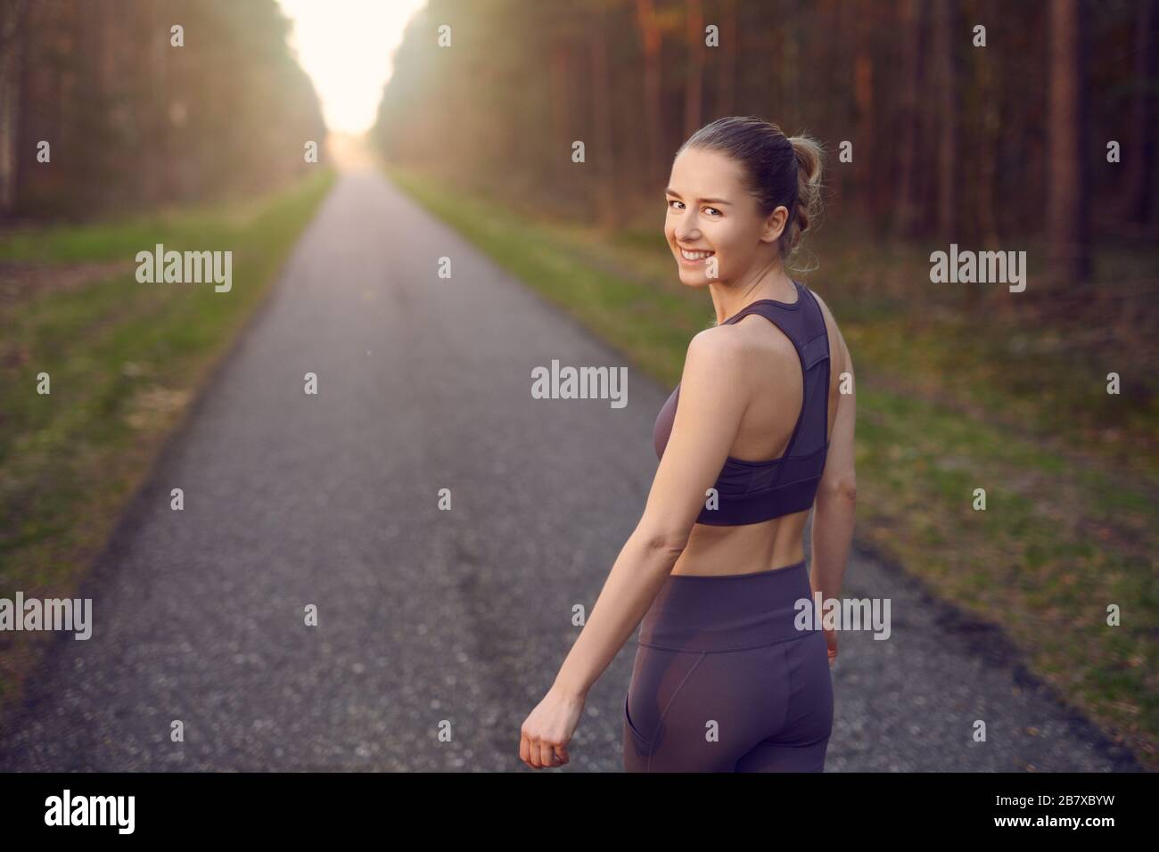 Die junge, athletische Frau passt beim Sonnenaufgang auf einer gerade sich zurückziehenden Straße durch Waldbäume und leuchtet am Ende der Sonne und lächelt Stockfoto