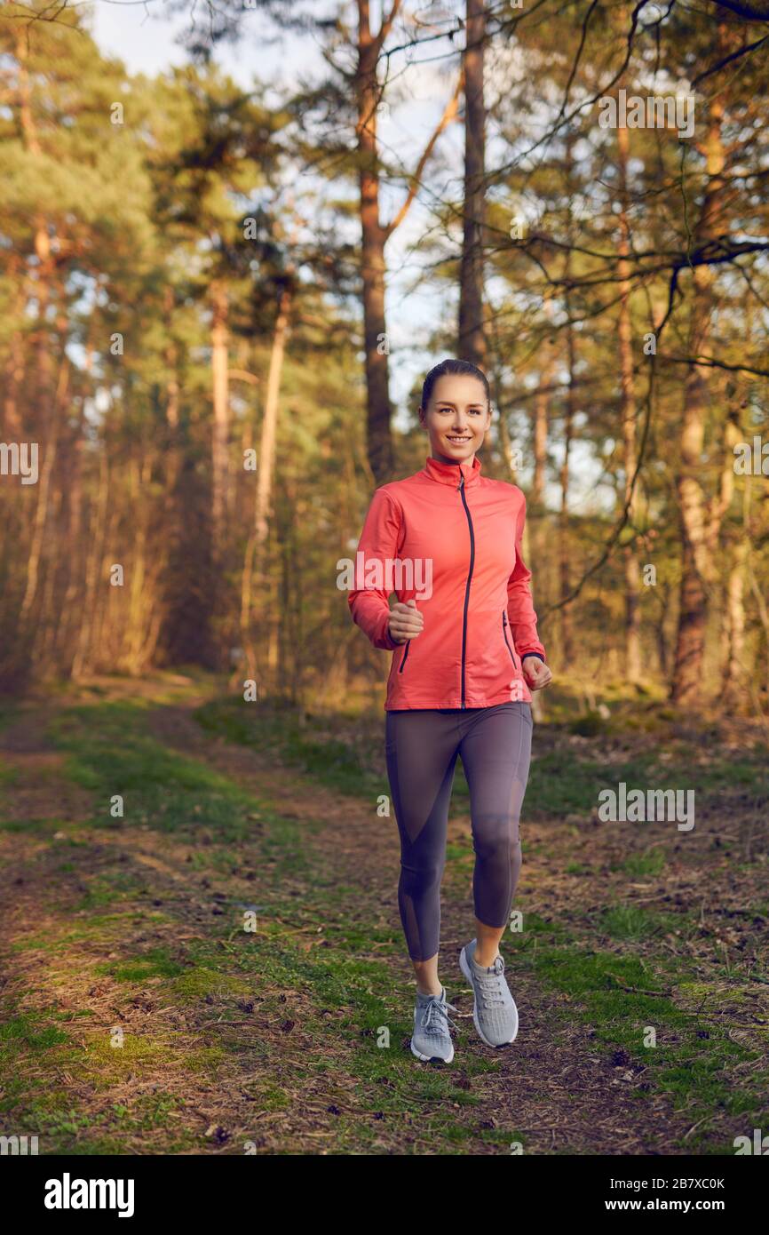 Gesunde Passform attraktive junge Frau Jogging im Wald entlang einer Waldbahn, die sich der Kamera mit einem fröhlichen Lächeln in einem aktiven Lebensstil nähert, Gesundheit a Stockfoto