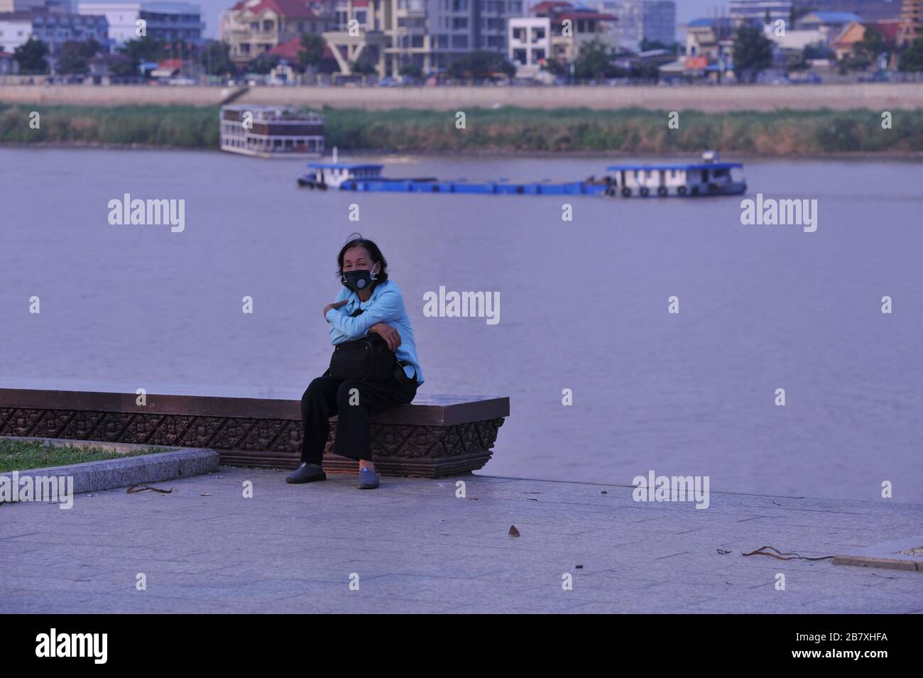 Eine ältere kambodische Frau in einer Gesichtsmaske sitzt während der Coronavirus-Pandemie allein am menschenleeren Flussufer. Phnom Penh, Kambodscha. © Kraig Lieb Stockfoto