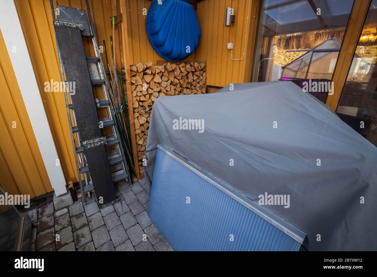 Blick auf den Hinterhof des Privathauses am Wintertag. Sommermöbel unter dem Deckel, Holzstapel auf gelbem Holzwandhintergrund. Stockfoto