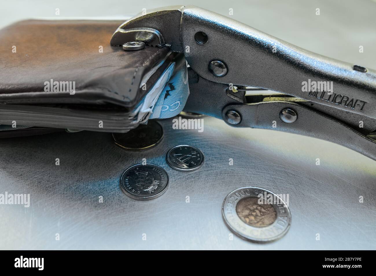 Auf einer Metalloberfläche wird eine Brieftasche mit einer Zange zusammengedrückt, auch einige (kanadische) Währungen sind umverteilt. Stockfoto